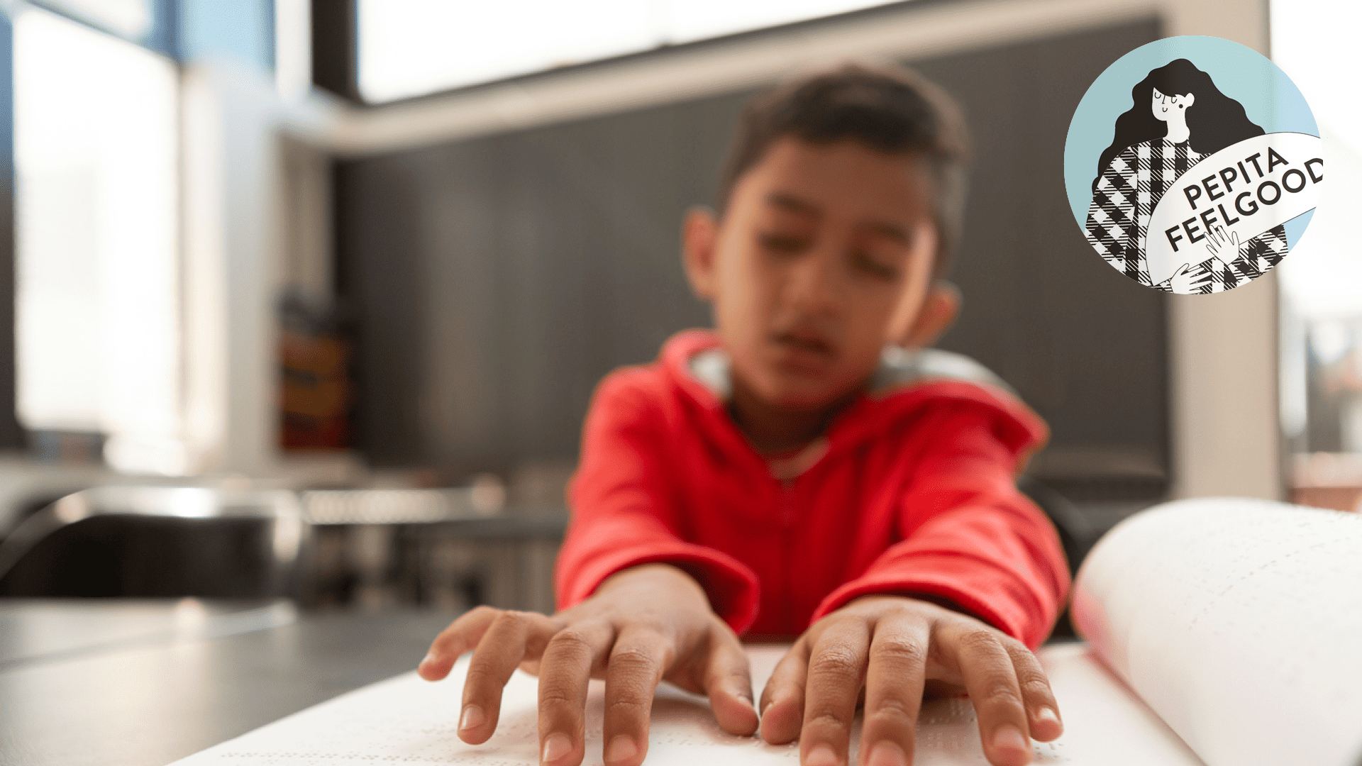 Ein Junge in einem roten Hoodie sitzt mit geschlossenen Augen an einem Tisch. Mit seinen Fingern erfühlt er Braille-Schrift in einem Buch.